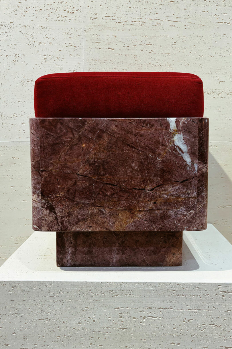 A deep red cushion and a solid, rectangular brown marble base, displayed on a white platform against a light stone wall.