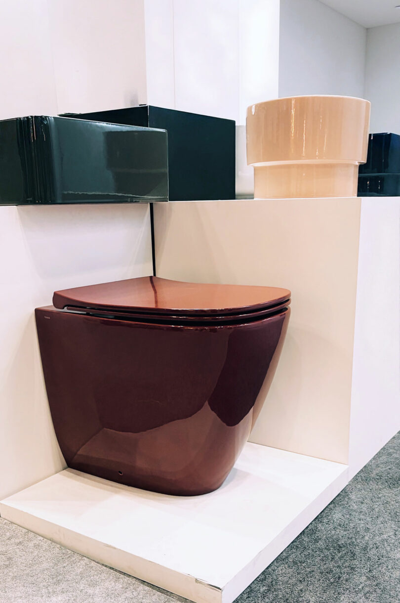 A modern maroon toilet displayed on a white platform, surrounded by other geometric bathroom fixtures in different colors.