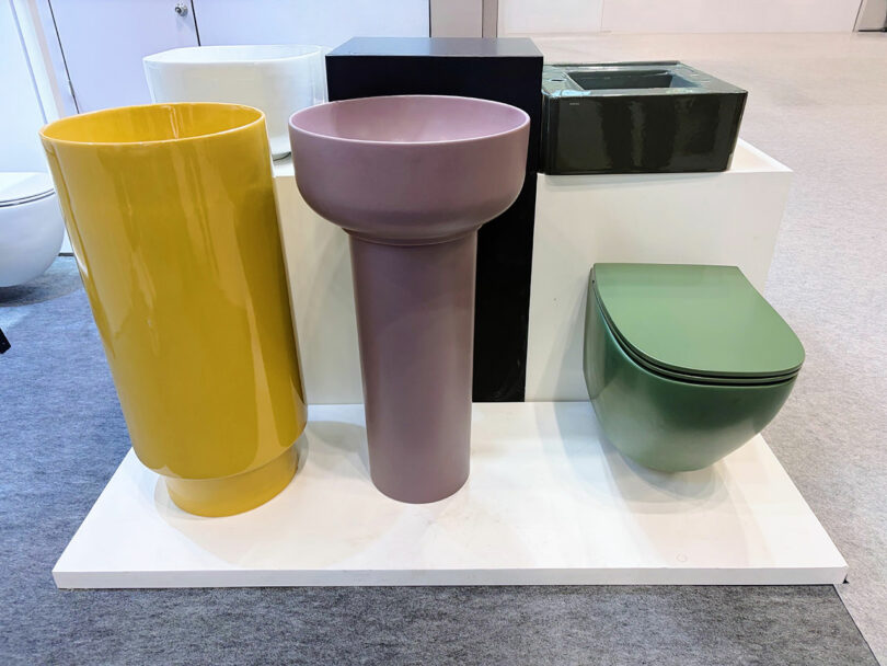 A display of modern bathroom fixtures—a yellow freestanding basin, a purple pedestal sink, a green wall-mounted toilet, and a sleek rectangular green sink.