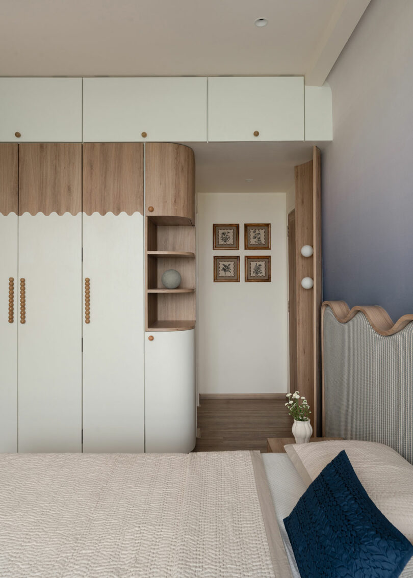 Dormitorio moderno con armario empotrado blanco y de madera, una cama beige con almohadas azules y tres fotografías enmarcadas en la pared del pasillo.