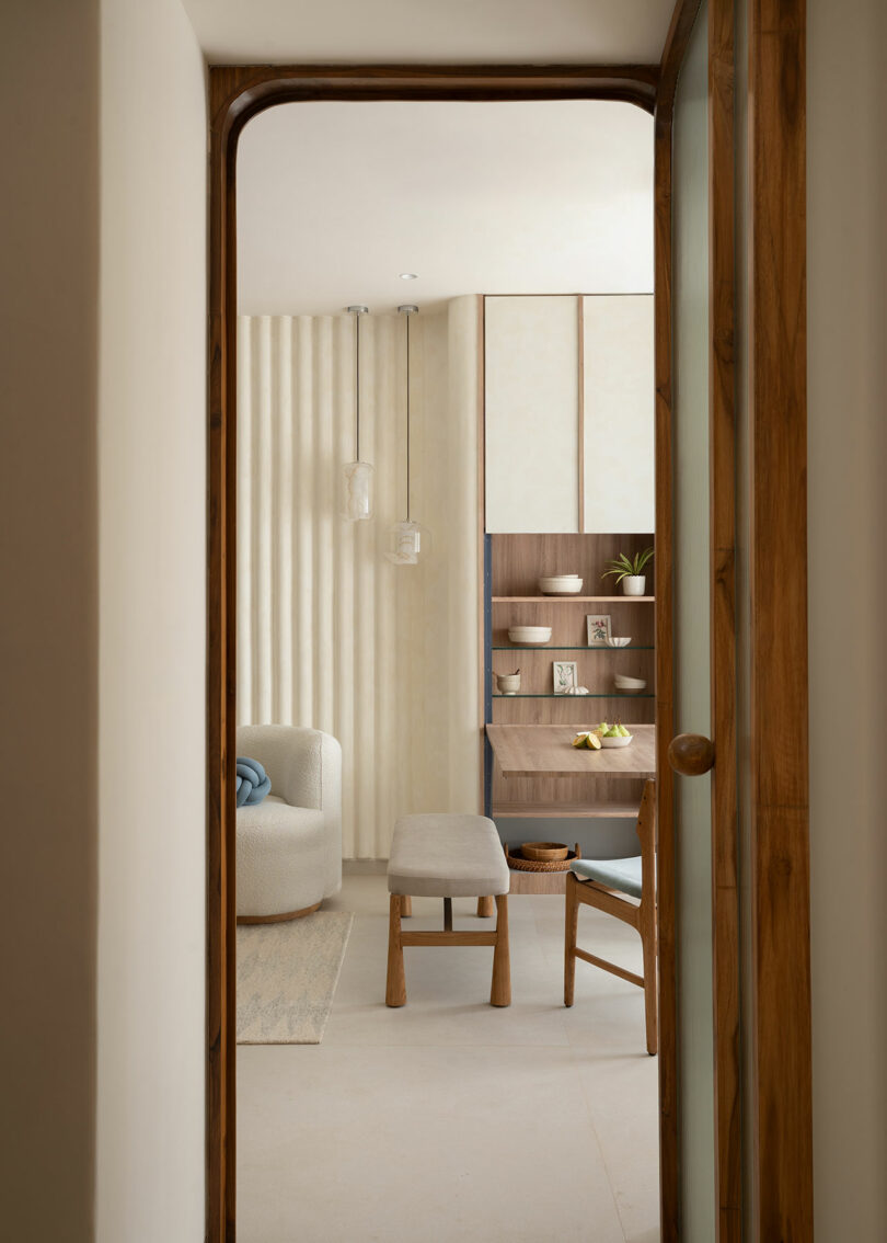 Una puerta revela una habitación minimalista con un sillón blanco curvo, taburetes de madera, sillas, estanterías repletas de elementos decorativos y paredes en tonos neutros.
