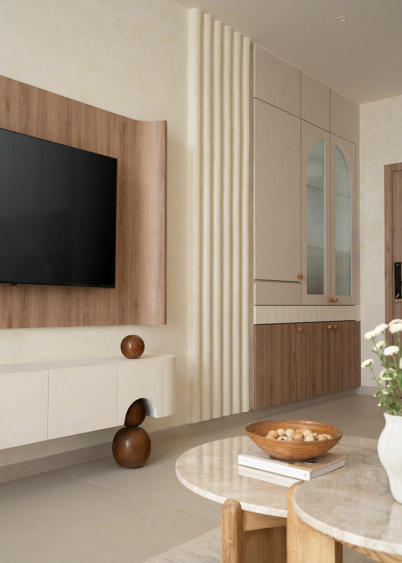 Sala de estar moderna con TV montada en la pared, gabinetes flotantes, detalles en madera, esferas decorativas y mesa de centro redonda con cuenco y jarrón. El trastero incorporado cuenta con una puerta de cristal arqueada.