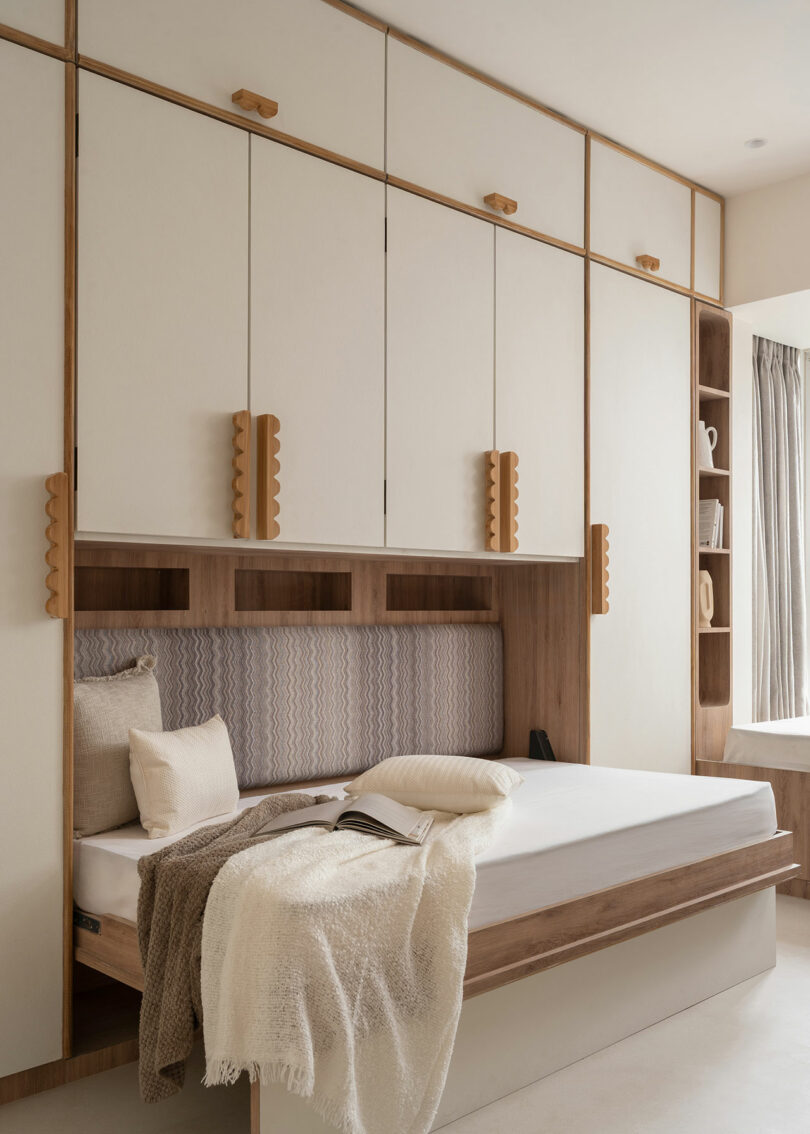 Dormitorio de estilo minimalista con cama con estructura de madera, ropa de cama neutra, armarios empotrados arriba y estantes a la derecha; La luz natural entra por la ventana con cortinas beige.