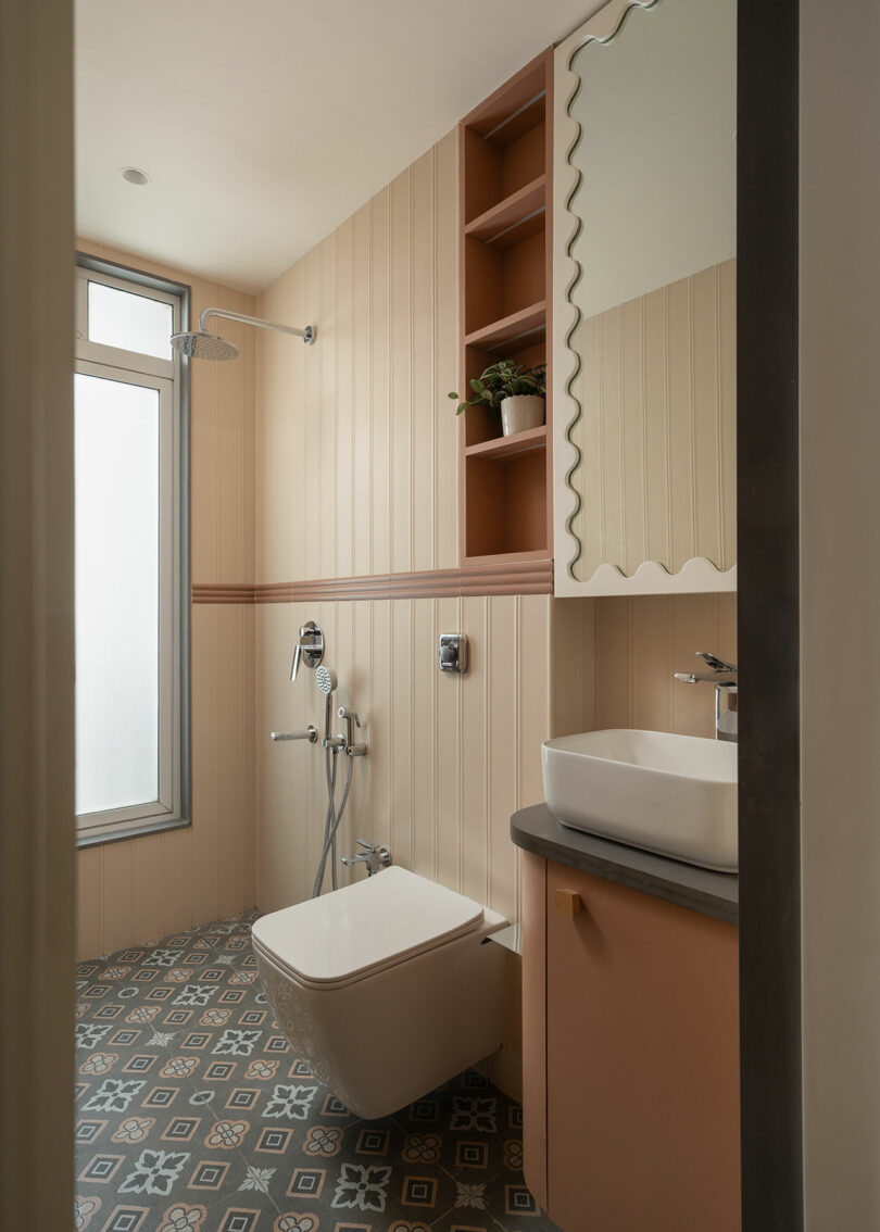Cuarto de baño moderno con suelo de baldosas estampadas, inodoro montado en la pared, ducha y lavabo con espejo rectangular. Los estantes incorporados contienen plantas y otros artículos. Las ventanas esmeriladas proporcionan luz natural.