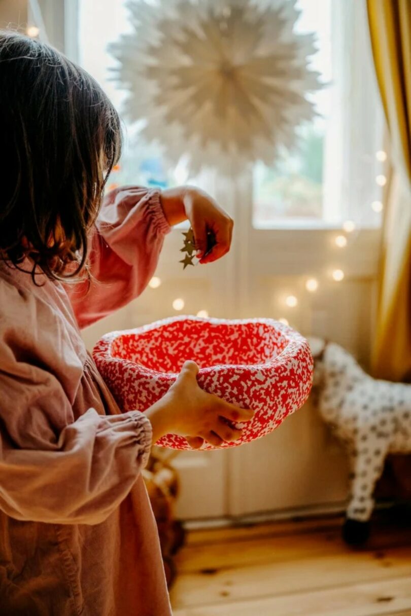 A child in a pink dress holds a red patterned bowl, dropping a small star ornament into it, surrounded by festive decorations and lights—perfect inspiration for heartfelt Valentine's Day gifts
