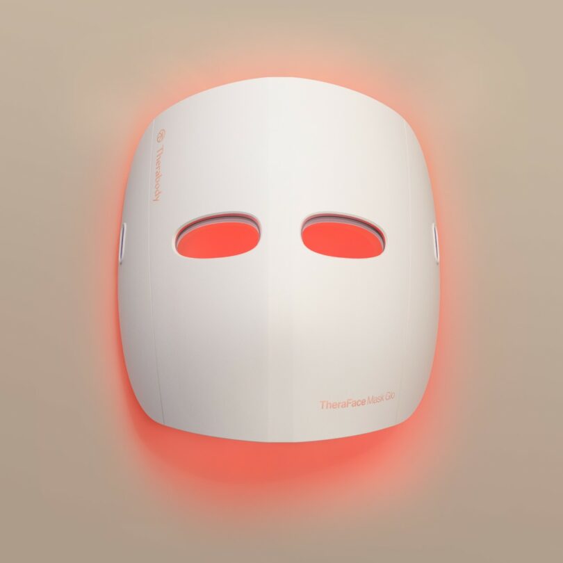 A white facial mask device with eye cutouts and glowing red light around its edges, displayed against a beige background