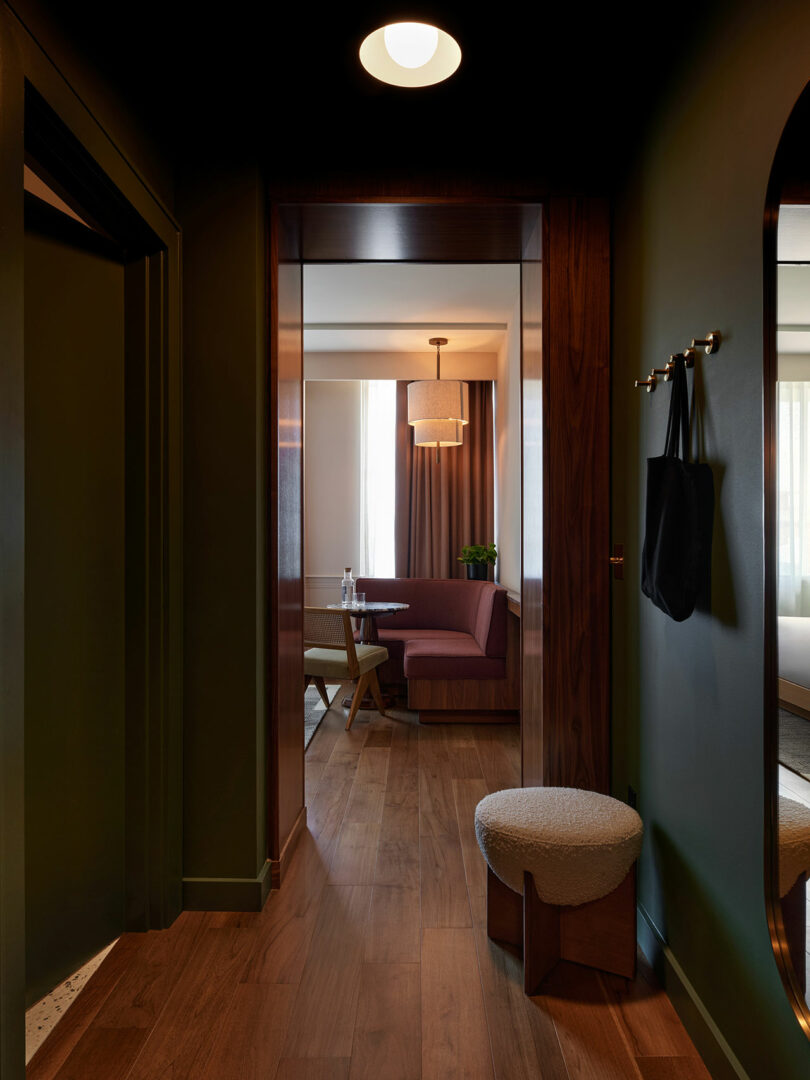 Hallway with dark green walls and wood floors exudes a Municipal Grand charm, featuring a round stool, coat hooks with a bag, and a view into a sitting area with a table, purple sofa, and large window draped in curtains.