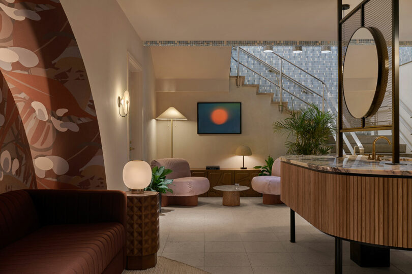 Modern lobby in the Municipal Grand features curved furniture, round light fixtures, indoor plants, a staircase with glass railing, abstract wall art, and patterned wallpaper on the left wall.
