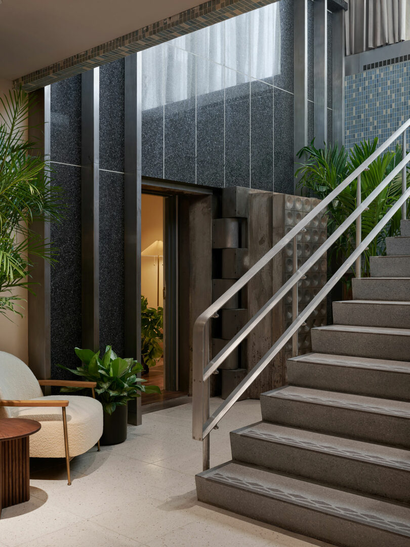 Modern indoor space at Municipal Grand with gray tiled stairs, metal handrail, green potted plants, a round chair, and a partially open door leading to a warmly lit room.