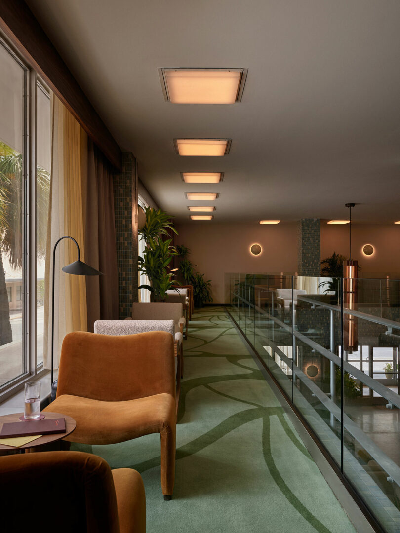 Modern lounge area at Municipal Grand with brown chairs, small tables, floor lamp, green carpet, and large windows. Glass railing overlooks the lower level; potted plants and ceiling lights line the spacious retreat.
