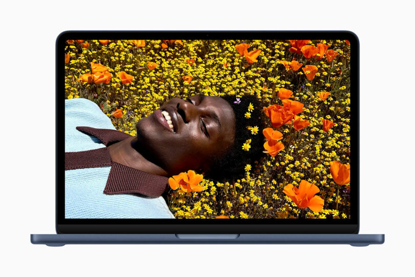 Una computadora portátil (posiblemente un dispositivo Apple) muestra una imagen de una persona tumbada en un campo de flores amarillas y naranjas, sonriendo y mirando hacia arriba.