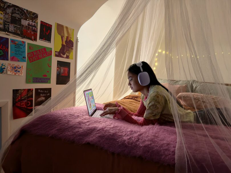 Una niña con audífonos yace en una cama cubierta con una manta rosa y usa una computadora portátil Apple bajo un dosel transparente, rodeada de carteles y luces de cadena.