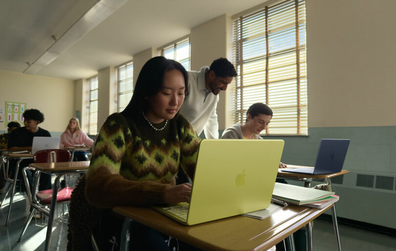 Un estudiante usa una computadora portátil Apple amarilla en un escritorio, mientras un maestro ayuda a otro estudiante en el aula y varios otros estudiantes trabajan.