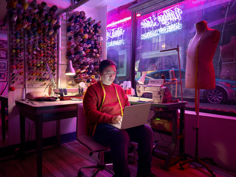 En un estudio de costura, un hombre se sienta en un escritorio sosteniendo una computadora portátil Apple, rodeado de carretes de hilo, máquinas de coser, lámparas y batas, con luces de neón brillando a través de las ventanas.