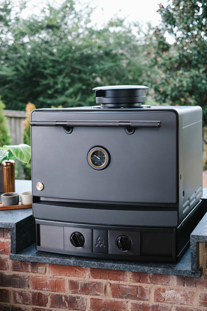 An Emberhaus black outdoor pizza oven with a built-in thermometer sits on a brick countertop in a cozy backyard setting.