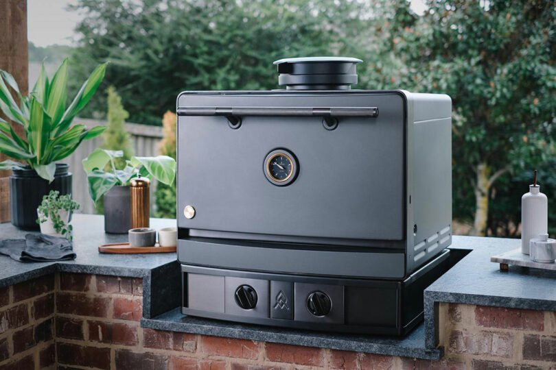 An Emberhaus black outdoor oven with a temperature gauge and control knobs sits on a brick counter, surrounded by plants and kitchen items.