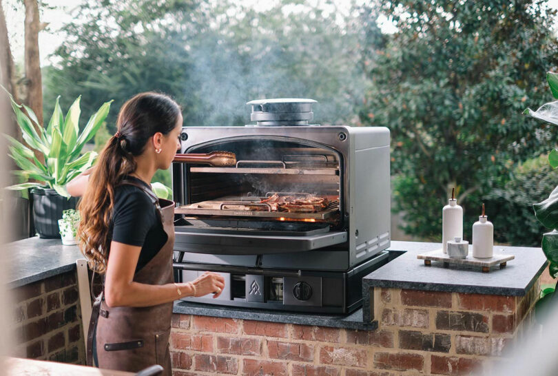 A woman in an apron cooks food on an outdoor brick kitchen using a large Emberhaus metal oven with visible smoke, surrounded by plants and condiments.