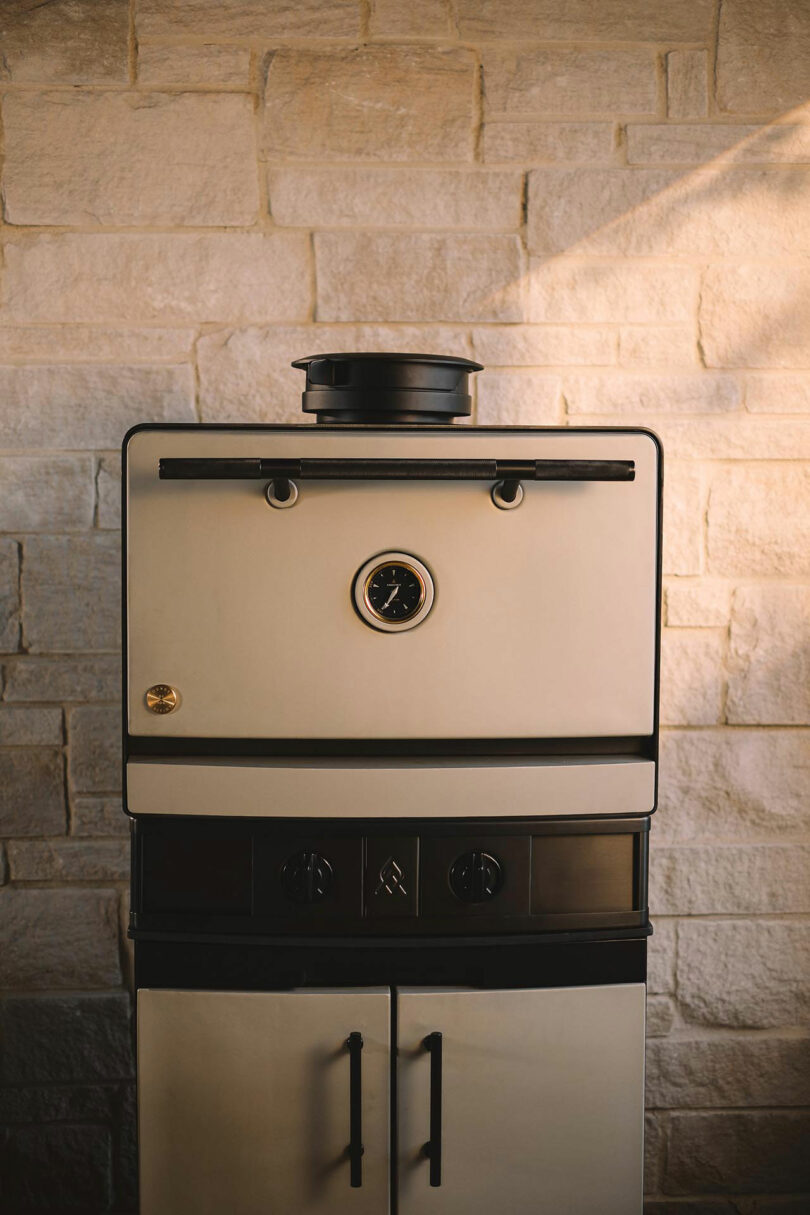 An Emberhaus vintage-style oven with a central temperature gauge sits against a light stone wall, sunlight casting a gentle shadow across the surface.