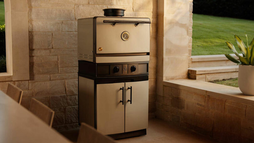 An Emberhaus stainless steel outdoor oven with double doors and control knobs is placed against a stone wall in a patio setting beside a potted plant.