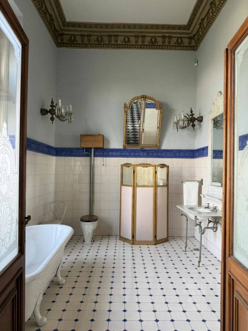 An ornate vintage bathroom by Coco Greenblum features a clawfoot tub, wooden toilet, folding screen, large mirror, and wall sconces on tiled floor and walls with blue trim.