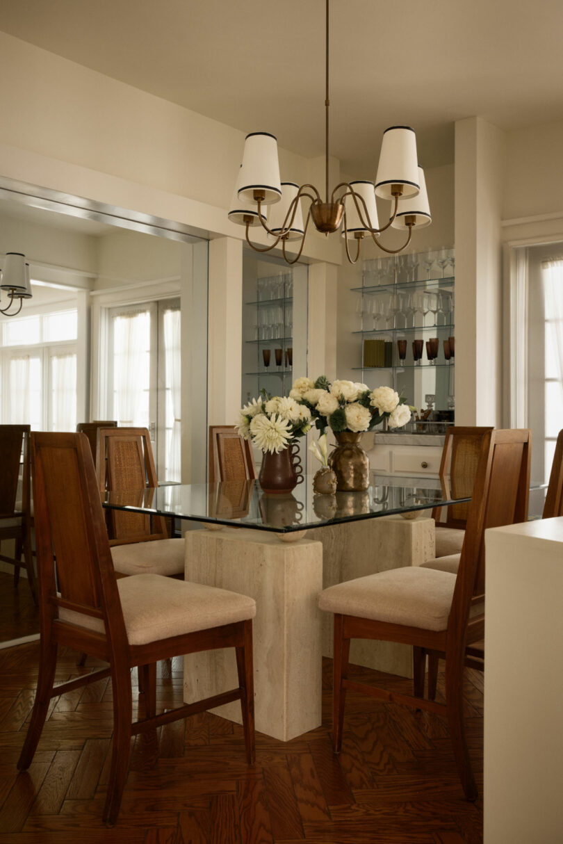 A glass-top dining table with six wooden chairs, a floral centerpiece, and a chandelier graces this Coco Greenblum-inspired room with mirrored walls and wood flooring.