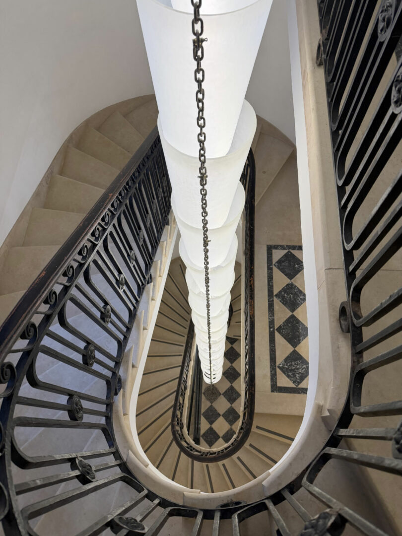 Spiral staircase with dark railings viewed from above, featuring a central hanging light fixture and a patterned tile floor at the bottom—a signature touch reminiscent of Coco Greenblum’s elegant design aesthetic.