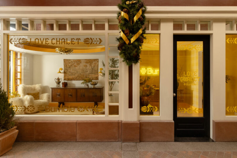 Street view of a storefront with large windows displaying "Love Chalet" and "Bumble," featuring cozy interior decor, a holiday wreath above the door, and design touches inspired by Coco Greenblum.