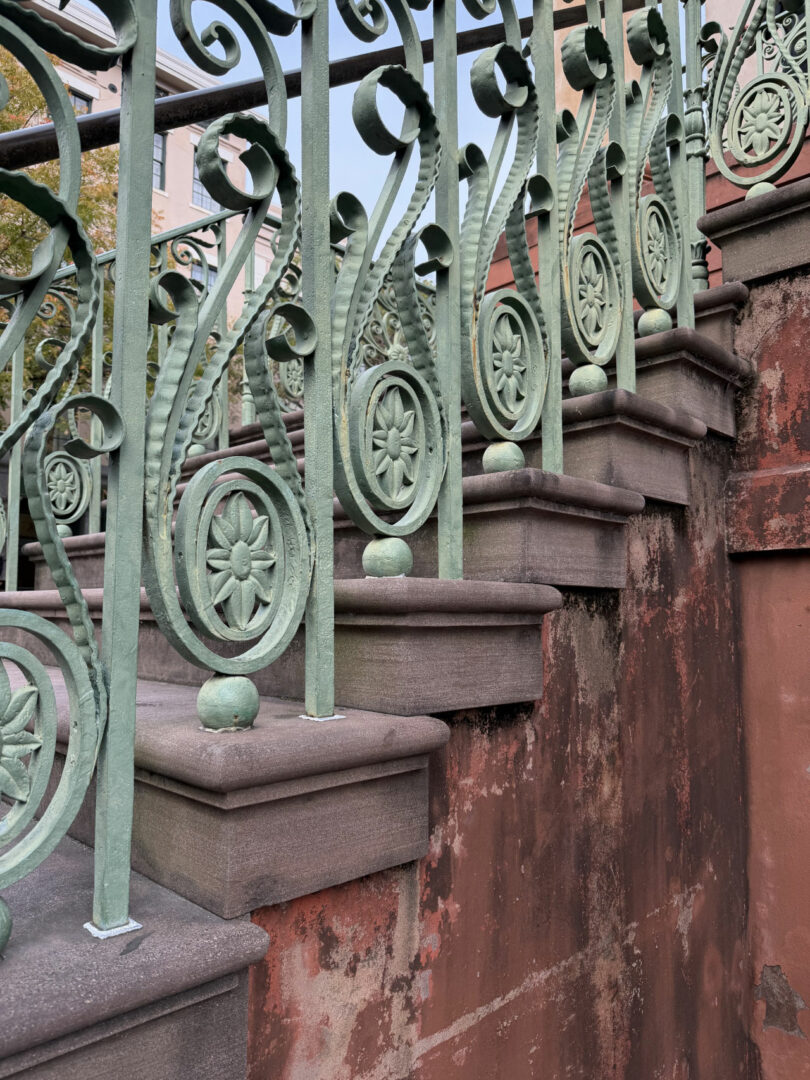 Green ornate metal railing with floral patterns, inspired by Coco Greenblum, lines a weathered stone staircase, which has red and brown discoloration on the wall side.
