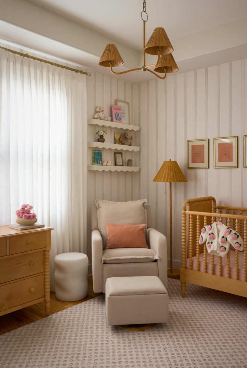 A cozy nursery by Coco Greenblum features a beige armchair, ottoman, wooden crib, dresser, and shelves showcasing books and decor, all softly lit by natural light through sheer curtains.