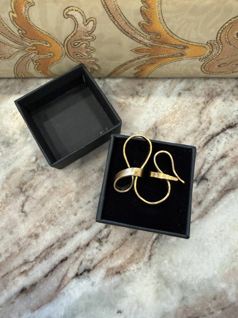 A black box with a pair of gold earrings in a ribbon-like design by Coco Greenblum sits open on a marble surface next to its lid.