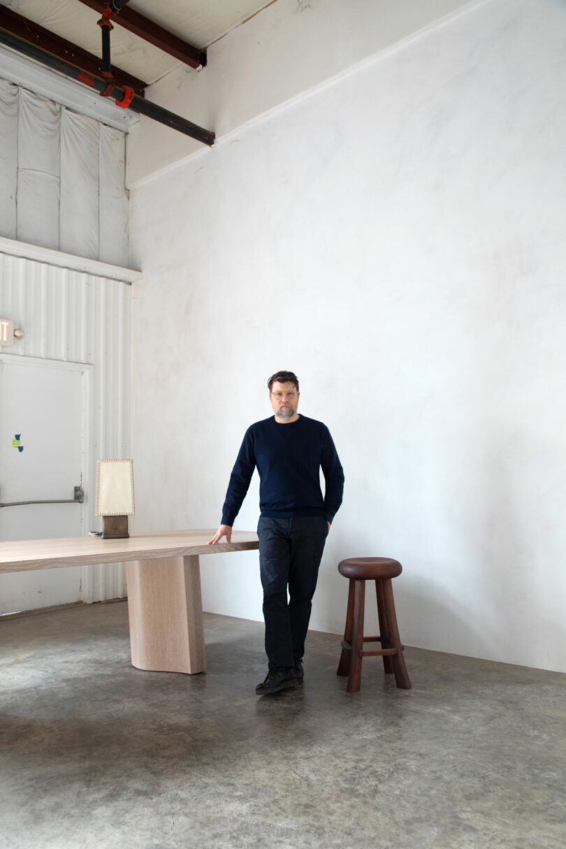 A man in a navy sweater stands in a minimalist room, leaning against a light wood table with a lamp—his relaxed pose echoing the effortless style of a Friday Five feature—next to a wooden stool on a concrete floor.