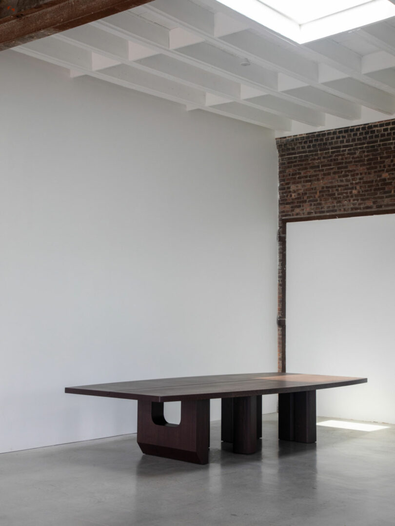 A large, dark wooden table with geometric legs stands in a minimalist room with white walls, a brick accent, and a skylight above—perfect for gathering the team for your next Friday Five discussion.