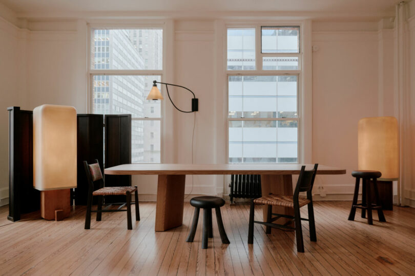 A minimalist office or dining room, perfect for a Friday Five gathering, features a large wooden table, four chairs, ambient floor lamps, and expansive windows framing a stunning cityscape.