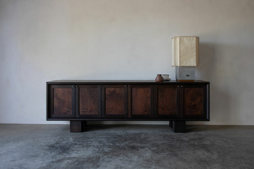A dark wooden sideboard with four cabinet doors, featured in our Friday Five, stands against a plain wall and holds a ceramic vase and a square lamp with a stitched-edged shade.