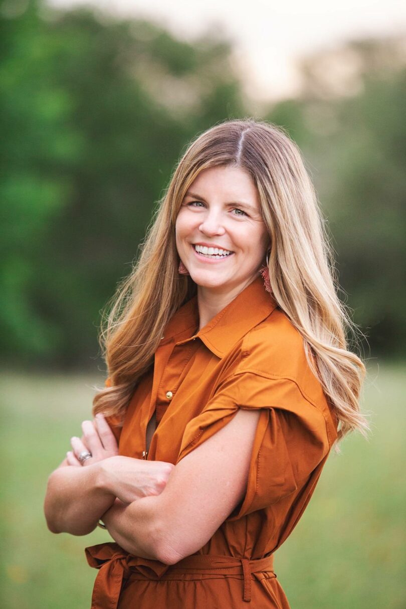 Una mujer con cabello largo y rubio y un vestido naranja estaba afuera con los brazos cruzados y sonreía a la cámara. Al fondo hay árboles verdes borrosos, que capturan el ambiente relajado del viernes 5.