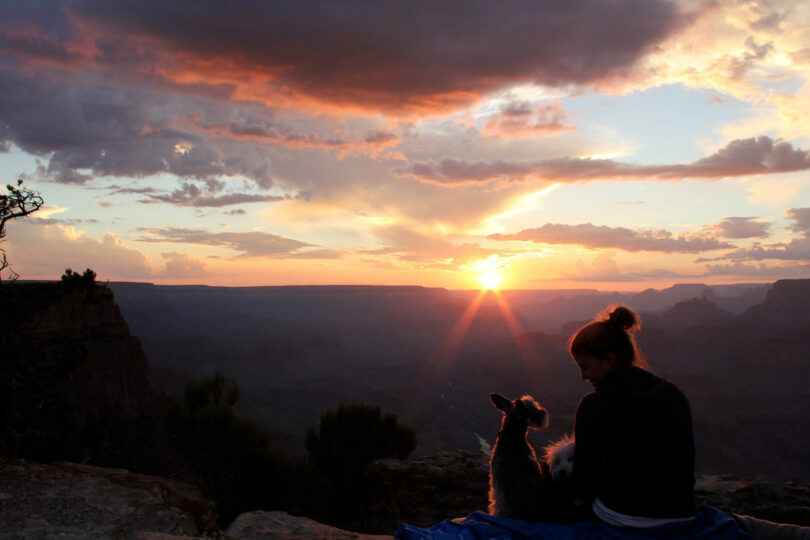 Un hombre y un perro se sientan sobre una manta, contemplando la puesta de sol sobre un cañón y las nubes flotando sobre los acantilados distantes: el momento perfecto del viernes 5 para relajarse y sumergirse en la belleza de la naturaleza.