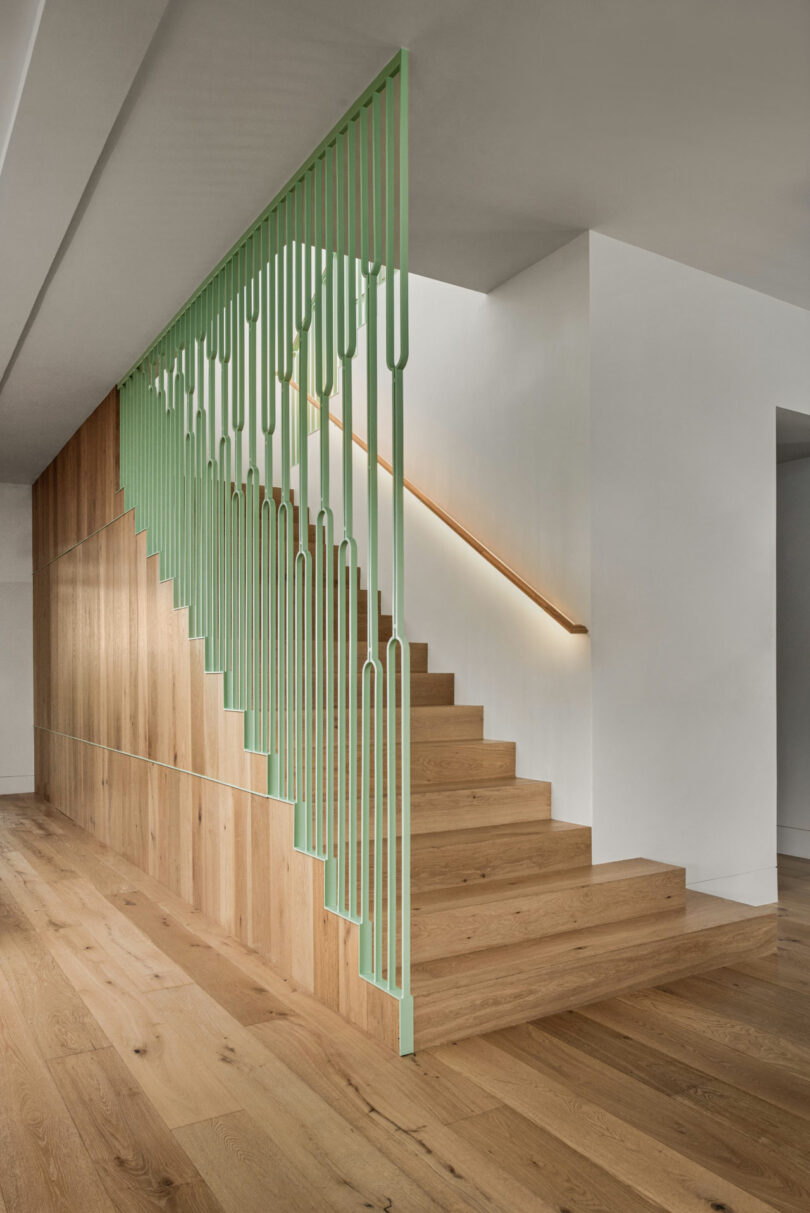 Una escalera moderna con escalones de madera y barandilla metálica geométrica de color verde claro forma parte de un interior minimalista estilo Friday 5th con paredes blancas y paneles de madera lisos.
