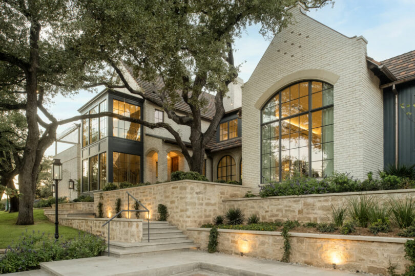 Casa moderna de dos pisos con ventanas grandes, exterior de ladrillo claro, muro de contención de piedra y patio ajardinado con árboles maduros, perfecta para tus reuniones de los viernes.