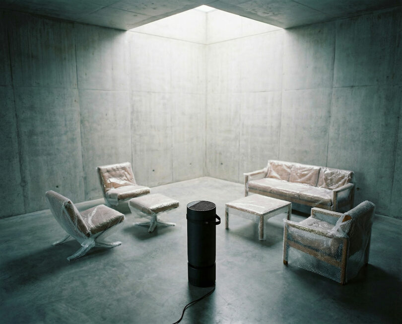 Una habitación de hormigón con un tragaluz contiene muebles envueltos en espuma rodeados de objetos cilíndricos negros en el suelo.