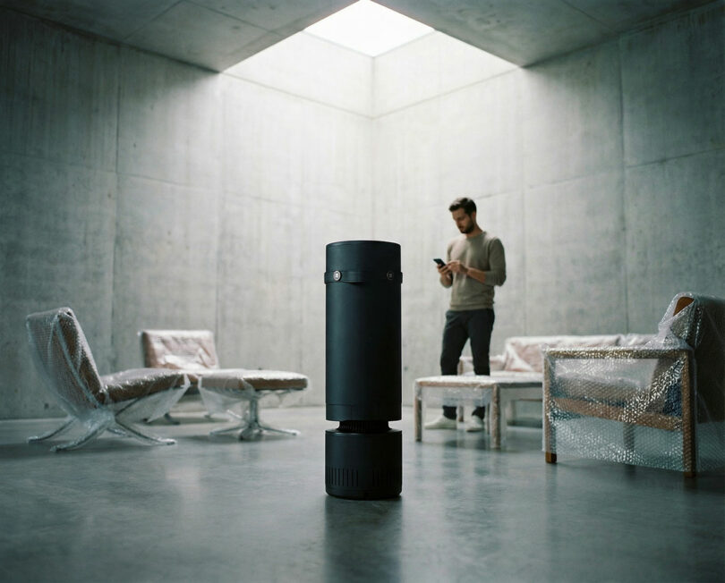 Una instalación cilíndrica negra se asienta sobre el piso de concreto de una habitación minimalista con paredes de concreto; un hombre se encuentra al fondo cerca de muebles envueltos en plástico de burbujas.