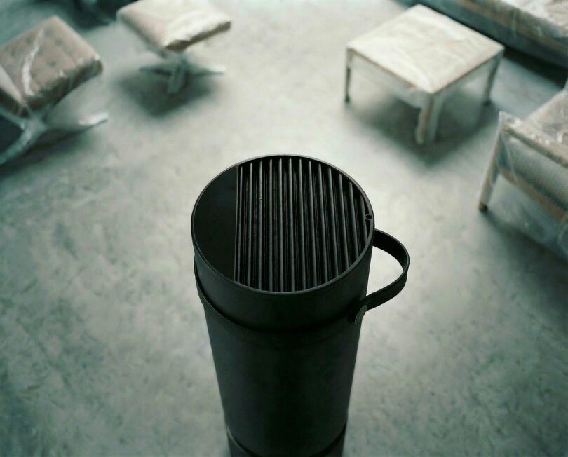 Primer plano de una estufa cilíndrica negra o de un calentador en una habitación con muebles envueltos en plástico y piso de concreto.