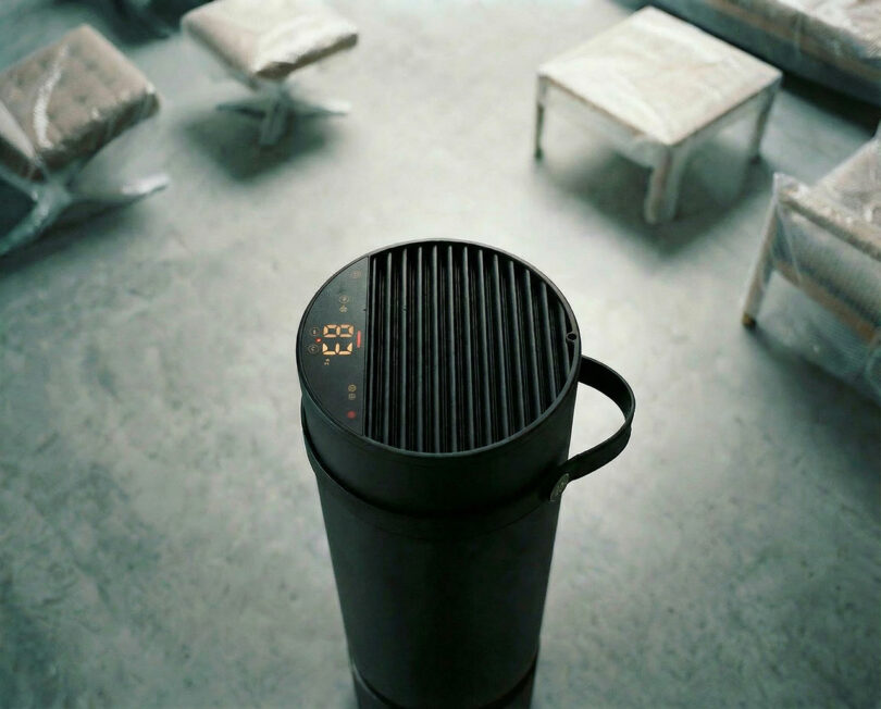 Hay un purificador de aire cilíndrico negro sobre el suelo de hormigón y se puede ver una envoltura de plástico cubriendo los muebles al fondo.