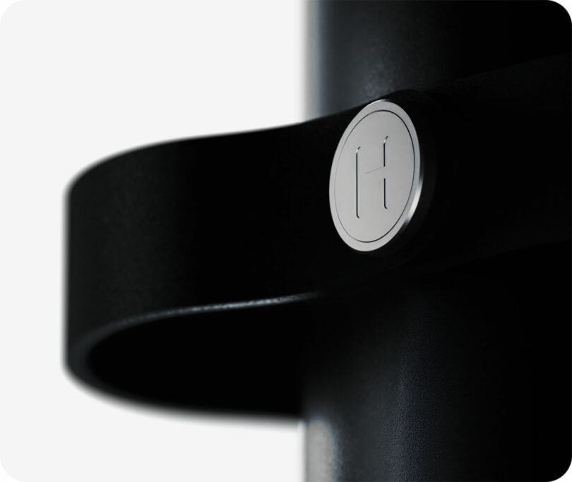 Primer plano de un objeto de metal negro con un botón plateado redondo con letras "h" grabado en él, sujeto a una correa curva negra sobre fondo blanco.