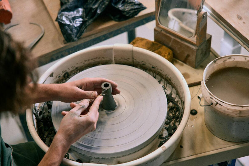 A person shapes a small clay object on a pottery wheel, using their hands and a tool, with Artek pottery materials and water nearby.