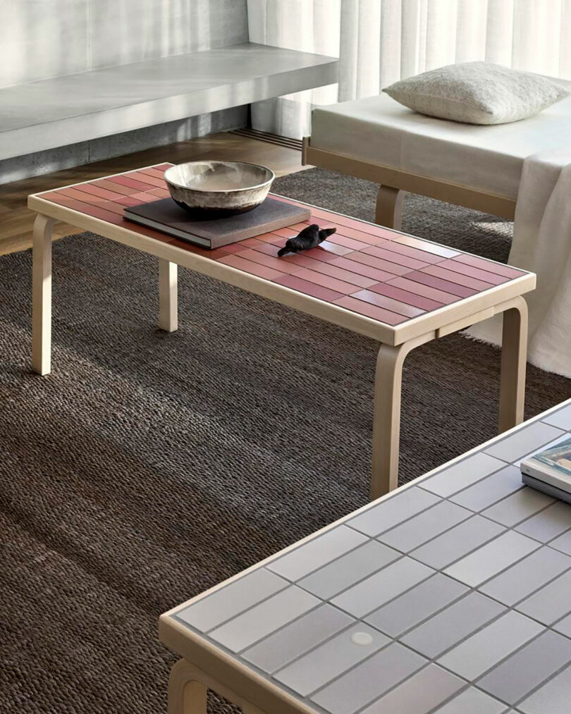 A rectangular Artek coffee table with a red tiled top holds a bowl, a book, and a small sculpture. The table stands on a dark rug next to a sofa and another tiled surface in this modern room.