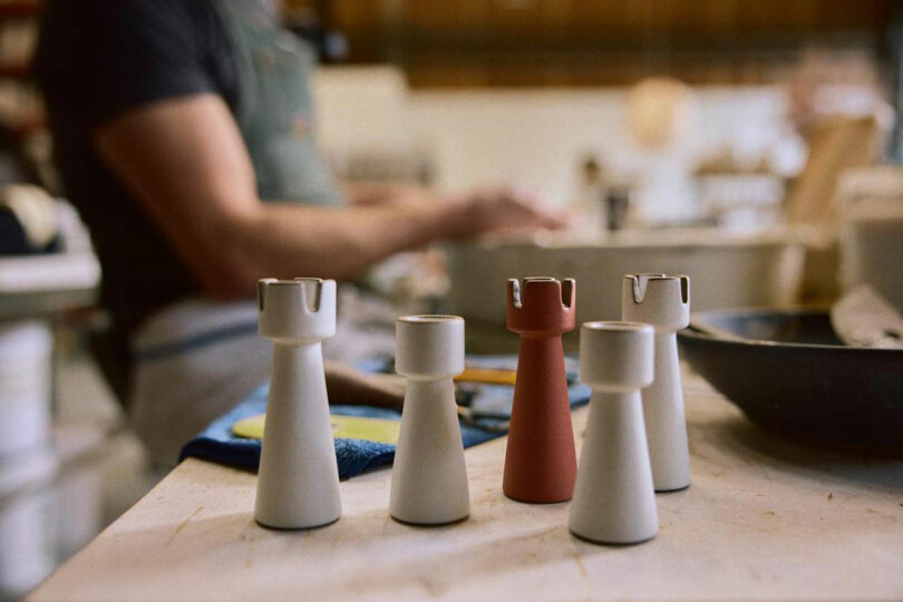 Five rook chess pieces, four white and one red, are arranged on a table at Artek, with a person working in the blurred background.