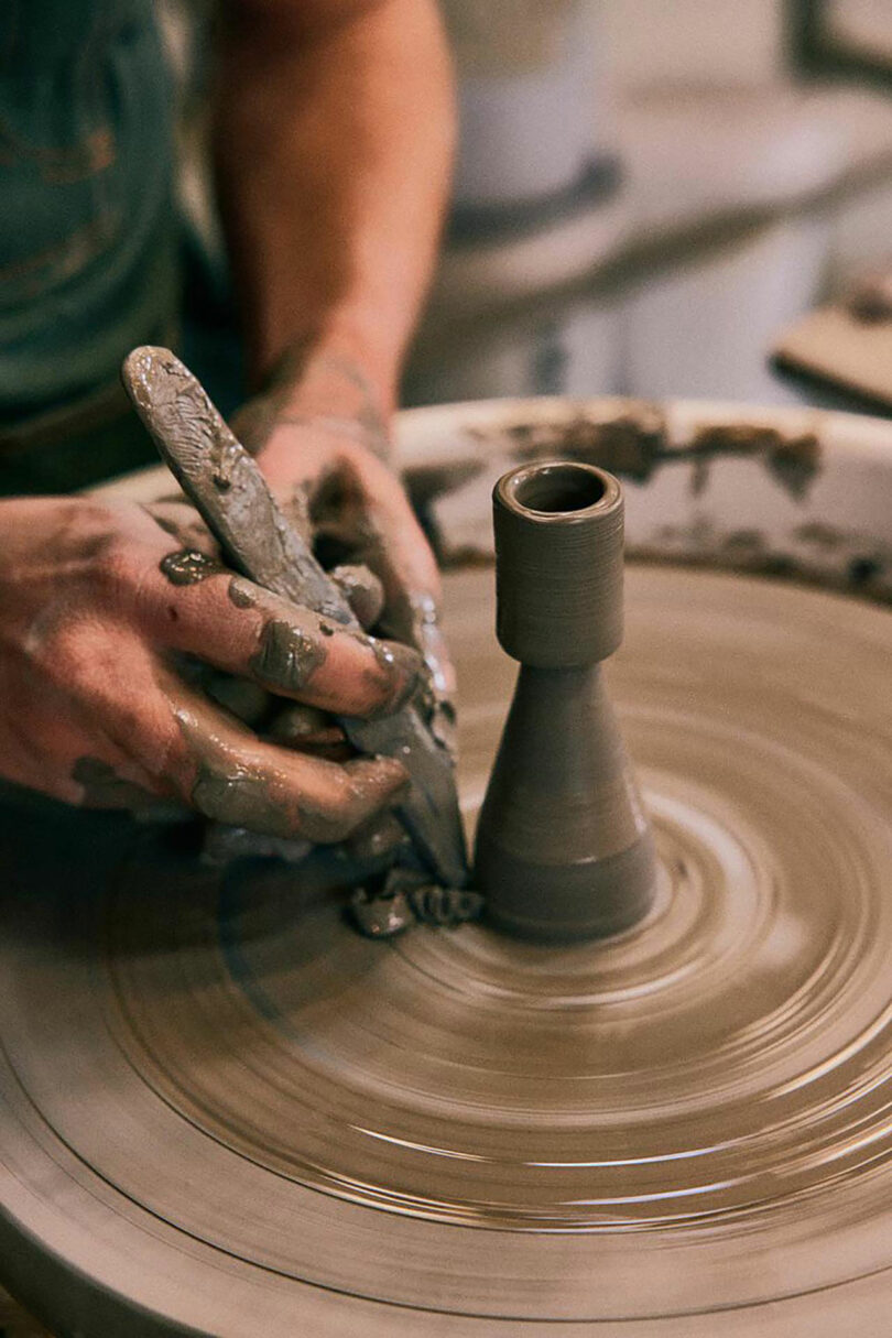 A person shapes wet clay on a pottery wheel, using their hands and a tool to form a narrow, cylindrical vase inspired by the minimalist design of Artek.