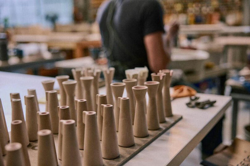 A row of unfinished ceramic vases sits on a worktable in a pottery studio, echoing the minimalist style of Artek, with a person working in the blurred background.