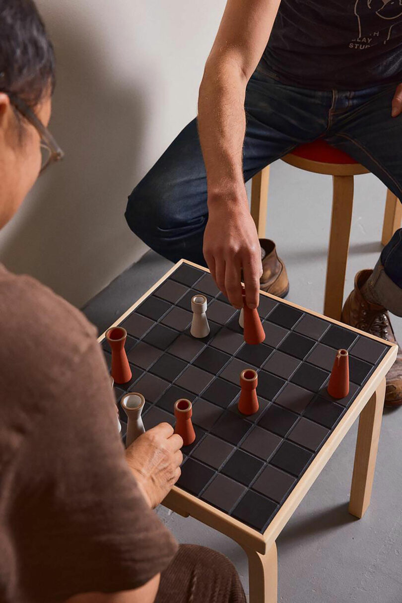 Two people play a board game with black squares and cone-shaped and cylindrical pieces on a small Artek wooden table.