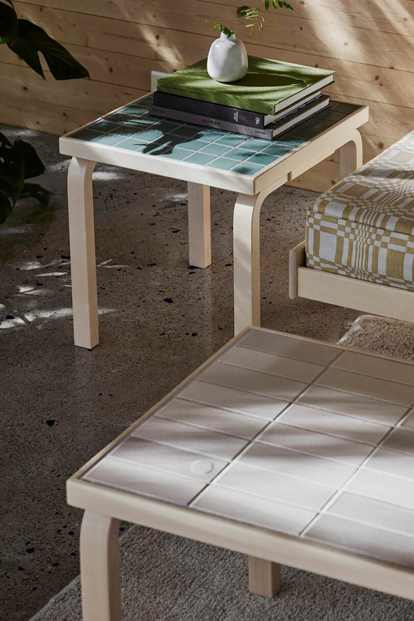 Two light wood Artek tables with tiled tops sit beside a cushioned bench; one table holds books and a small white vase, while sunlight casts gentle shadows across the surfaces.